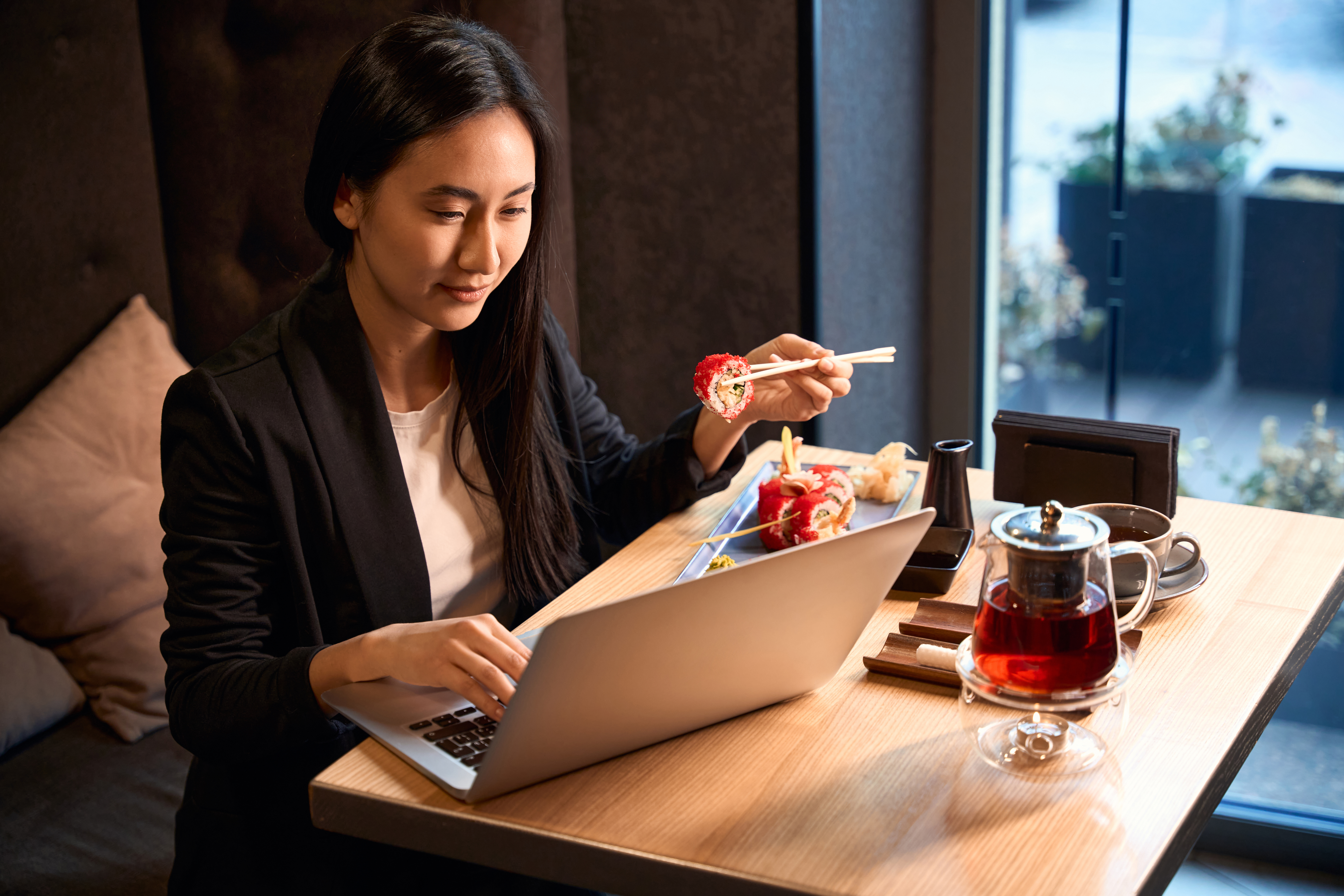 Asian woman eating sushi, drinking tea and checking project on laptop